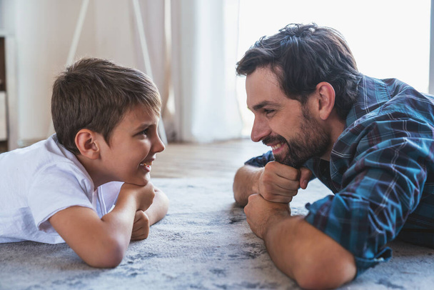 Portret van een kleine jongen en zijn vader die op de vloer liggen en glimlachen terwijl ze thuis naar elkaar kijken. - Foto, afbeelding