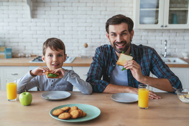 Muotokuva pieni poika ja hänen isänsä istuu pöydässä aamiaisen aikana kotona keittiössä. - Valokuva, kuva