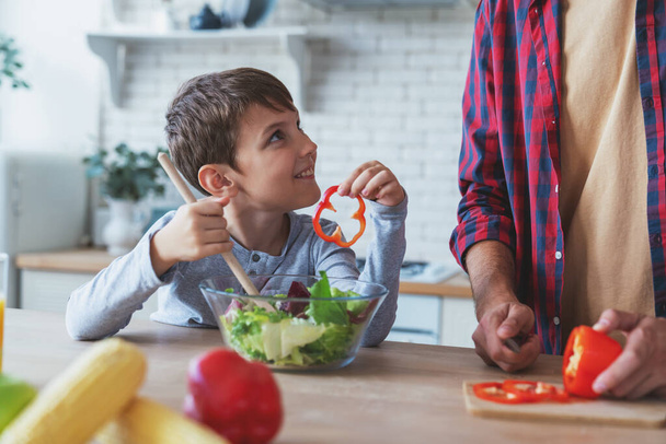 Cheerful father and his little son help each other while preparing a salad sitting at a table at home in the kitchen. - Photo, image