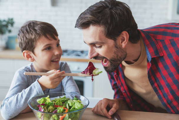 Cheerful father and his little son help each other while preparing a salad sitting at a table at home in the kitchen. - Photo, Image