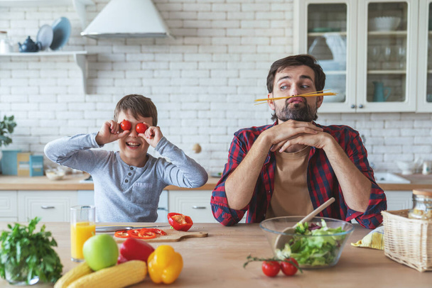 Alegre padre y su pequeño hijo se ayudan mutuamente mientras preparan una ensalada sentada en una mesa en casa en la cocina. - Foto, imagen