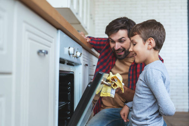 Veselý otec a jeho malý syn nahlédnout do trouby, zatímco vaří dort v jejich domácí kuchyni. - Fotografie, Obrázek