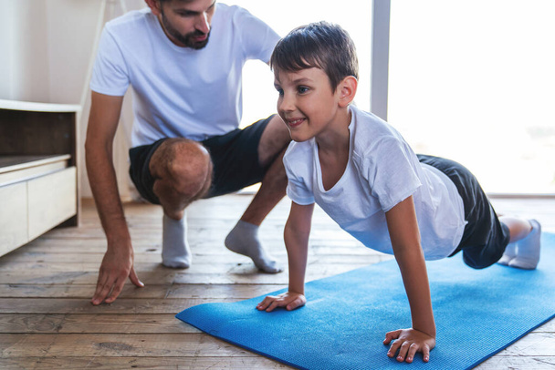Ένας νεαρός άνδρας βοηθά το γιο του κατά τη διάρκεια μιας γυμναστικής κάνοντας ασκήσεις στο σπίτι στο σαλόνι. - Φωτογραφία, εικόνα