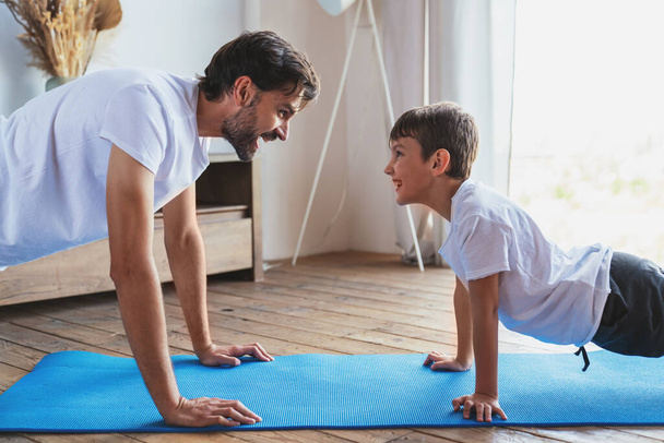 Um jovem ajuda seu filho durante um treino de fitness, fazendo exercícios em casa na sala de estar. - Foto, Imagem