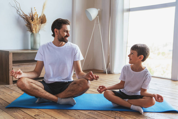 Młody mężczyzna pomaga synowi podczas treningu fitness uprawiając jogę w domu w salonie. - Zdjęcie, obraz
