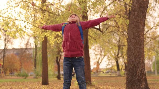 malá šťastná dívka s batohem hází listy vysoko nahoru a vesele se směje, dítě si hraje v podzimním parku, dítě procházky v městském parku, zvednout tvář při pohledu na zlaté listí podzim, podzimní sezóna - Fotografie, Obrázek