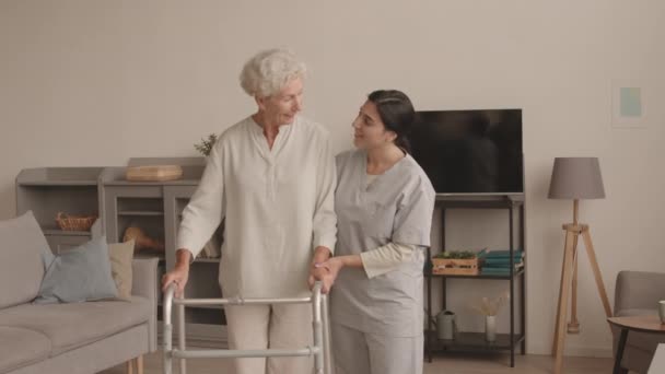 Medium steek schot van jonge vrouwelijke maatschappelijk werkster helpen senior vrouw met wandelaars ze glimlachen op camera poseren binnen in gezellig appartement - Video