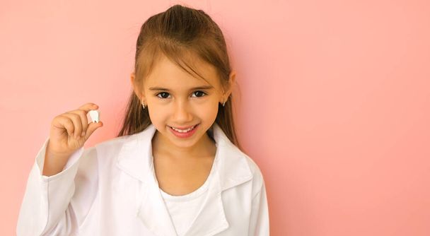 Cheerful girl portraying a doctor in a white medical coat holds a tooth in hand. Dental concept. Healthy and strong snow-white teeth from nature. Dental care and hygiene. Banner. Copy space - Photo, Image