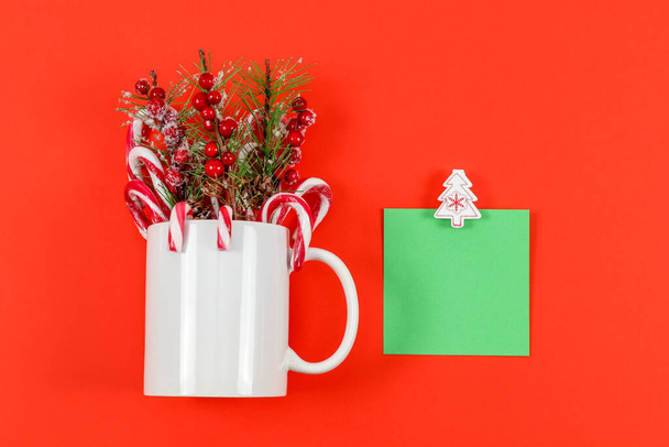 White porcelain mug with fir branches with red berries, candy lollipops and a green sheet of paper with a clothespin Christmas tree lie in the middle on a red background with space for text, top view close-up. - Zdjęcie, obraz