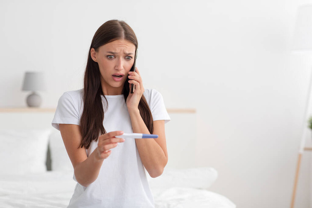 Shocked young european woman looks at pregnancy test, feels worried and calls her husband with news - Foto, Imagen