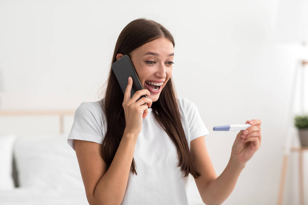 Cheerful excited pretty millennial european woman hold pregnancy test in bedroom interior and calling to boyfriend - Photo, Image