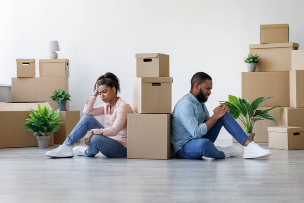infelice giovane ragazzo nero e signora sono stanchi e seduti sul pavimento in appartamento con scatole - Foto, immagini