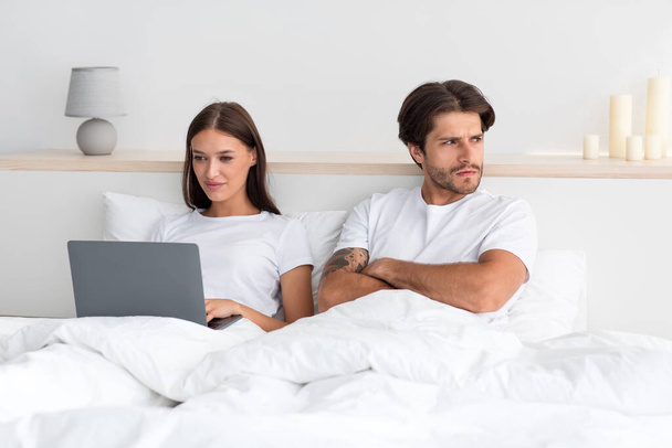 Junge Europäerin arbeitet am Laptop im Bett und ignoriert den verärgerten Mann mit verschränkten Armen, der im Schlafzimmer zur Seite schaut - Foto, Bild