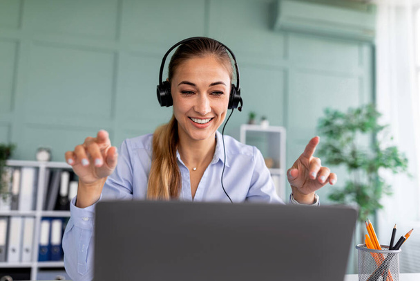 Mujer feliz en auriculares mirando a la computadora portátil y hablando durante el webinar, consultando al cliente o colega de negocios - Foto, imagen