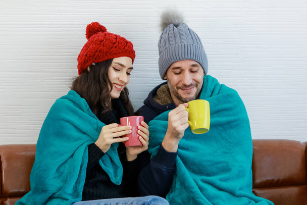 Happy caucasian lovers couple in wool sweater jacket knitted hat sit smile talk on couch cover by blanket together hold hot drink mug in hands in cold weather winter. - Photo, Image