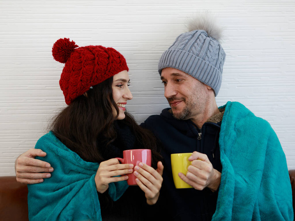 Romantic concept shot of caucasian lover couple in wool sweater jacket knitted hat cover and wrap by blanket together hold hot drink mug in hands in winter while husband hug wife with love. - Photo, Image