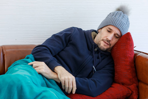 Caucasian sick unwell bearded man in wool jacket knitted hat lay down on leather sofa and pillow in living room wrap up by blanket having cold fever and headache in coronavirus outbreak pandemic. - Foto, Imagen