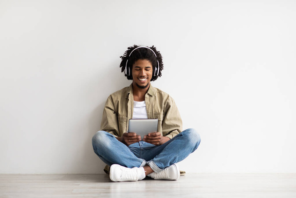 Młody czarny facet w słuchawkach bezprzewodowych Relaks na podłodze z tabletem cyfrowym - Zdjęcie, obraz