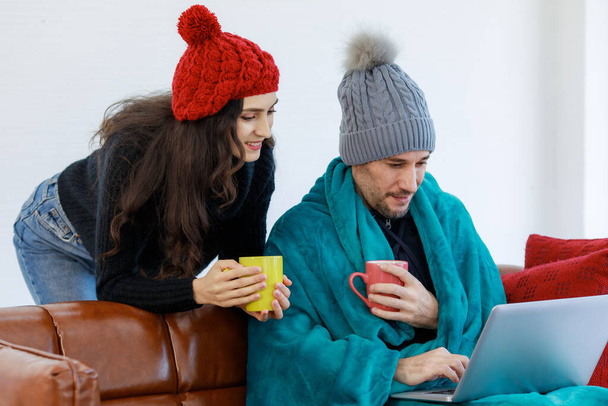 Kaukasisches Liebespaar in Wollpullover-Jacke und Strickmütze, bedeckt von einer warmen Decke, trinkt Heißgetränk und surft im Winter zu Hause gemeinsam im Internet einkaufen. - Foto, Bild