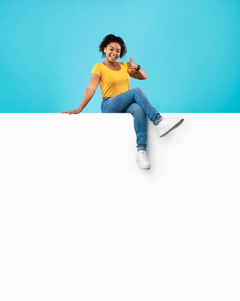 Sonriente mujer afroamericana sentada en un póster vacío, mostrando un gesto de pulgar hacia arriba en el fondo del estudio azul, maqueta - Foto, imagen