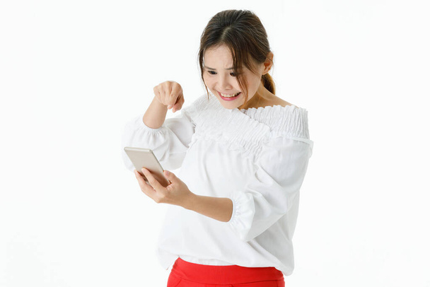 Geïsoleerde studio shot van Aziatische gelukkig bevredigen opgewonden vrouwelijke model in casual shirt met lange mouwen staande schreeuwen houden vuist omhoog vieren overwinning kijken naar smartphone op witte achtergrond. - Foto, afbeelding