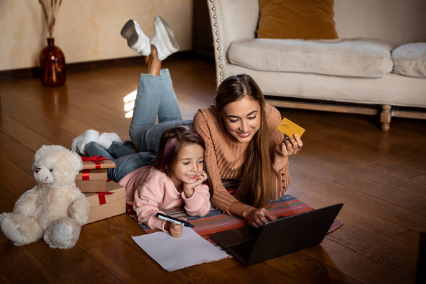 Mädchen schreibt Wunschliste, glückliche Mutter benutzt PC und Kreditkarte - Foto, Bild