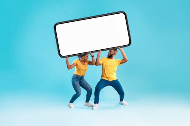Millennial black couple holding huge heavy cellphone with mockup on screen above their heads, space for mobile app - 写真・画像