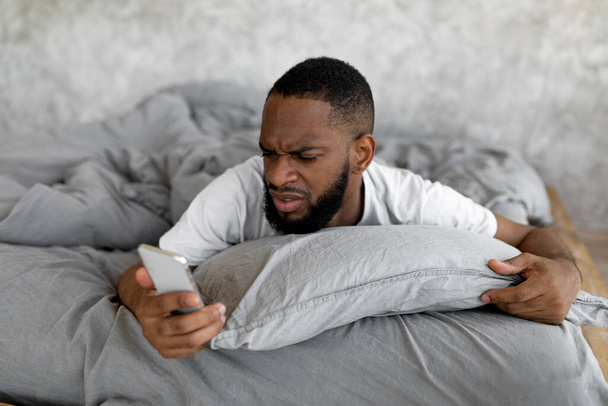 Confused black man looking at phone in bed - Fotó, kép