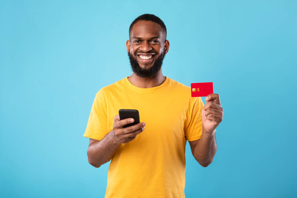 Fröhlicher junger schwarzer Mann mit Kreditkarte, der sein Handy für Online-Einkäufe, Bankgeschäfte oder elektronische Zahlungen benutzt - Foto, Bild