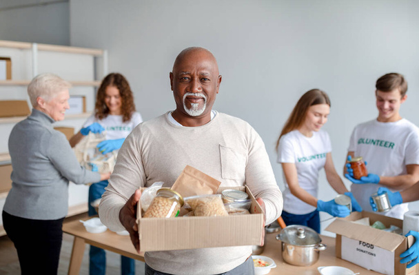 Centrum charytatywne. Szczęśliwy czarny starszy mężczyzna trzymając pudełko z darowizn jedzenie, patrząc i uśmiechając się do kamery - Zdjęcie, obraz