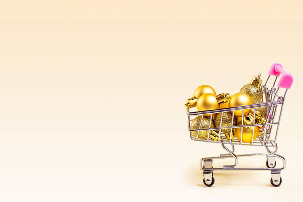 コピースペース付きの光の背景に金色のクリスマスボールを持つトロリーショッピングカート。クリスマスセール、割引、休日の買い物やプレゼントのコンセプト. - 写真・画像