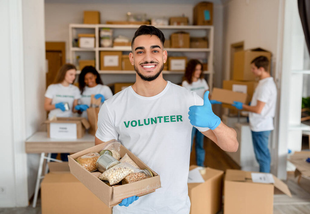 Hombre árabe feliz en el centro de voluntarios sosteniendo donaciones caja de papel, mostrando el pulgar hacia arriba y sonriendo a la cámara - Foto, Imagen