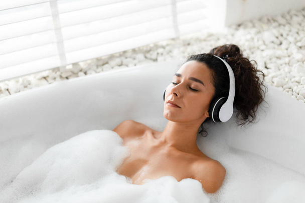 Ontspannen jonge vrouw luisteren naar kalmerende muziek in draadloze hoofdtelefoon, liggend in bubbelbad thuis, kopieer ruimte - Foto, afbeelding