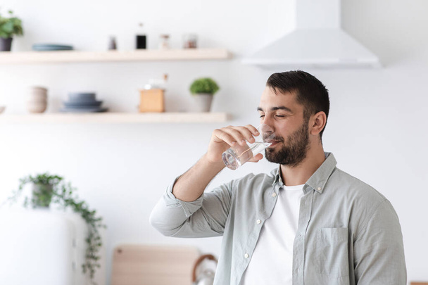 Europäischer attraktiver Mann mittleren Alters mit Bart trinkt Wasser aus Glas auf minimalistischem Kücheninterieur - Foto, Bild