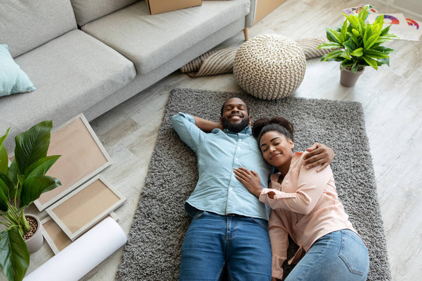 Ικανοποιημένη νεαρή αφροαμερικάνικη οικογένεια κείτεται στο πάτωμα, ξεκουραζόμενη μετά τη μετακόμισή της σε νέο διαμέρισμα στο σαλόνι - Φωτογραφία, εικόνα