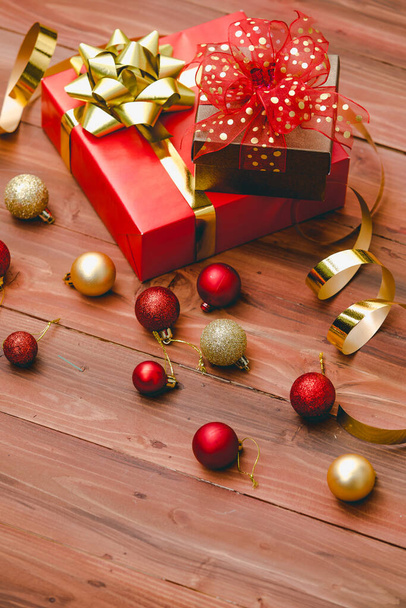 Close up studio shot of small brown present gift with gold dot on red ribbon bow tie placed on big paper wrapped box on dark brown pattern wooden table with shiny glossy Xmas decorative sphere balls. - Photo, Image