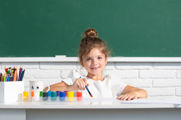 Painting school lesson, drawing art. Child girl draws in classroom sitting at a table, having fun on school blackboard background. - Photo, image