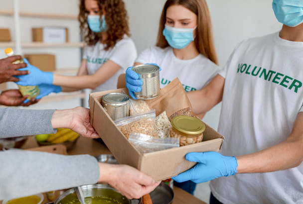 Молодые волонтеры в масках упаковывают еду в банки, банки и пакеты в коробки для пожертвований для бедных людей - Фото, изображение