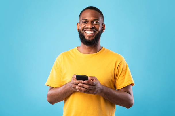 Joyeux jeune homme noir utilisant un smartphone, bavarder avec un ami, surfer sur Internet, avoir des leçons en ligne ou une réunion - Photo, image