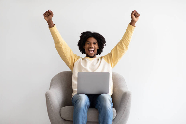 Online Wygraj. Szczęśliwy czarny facet świętuje sukces z laptopem, siedzi w fotelu - Zdjęcie, obraz