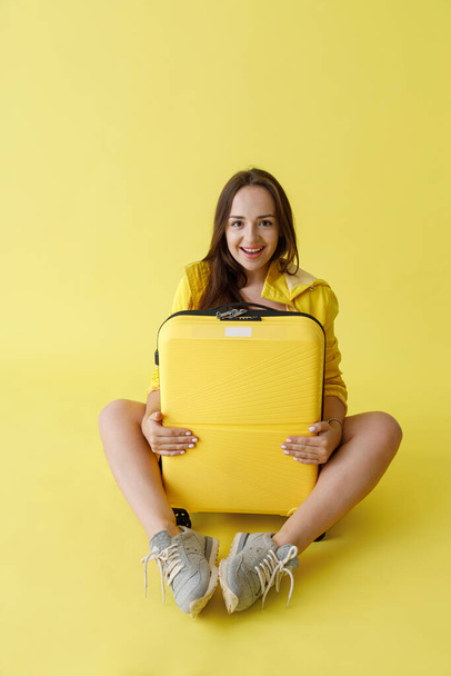 Radostná mladá žena s cestovní taškou sedí ve studiu - Fotografie, Obrázek