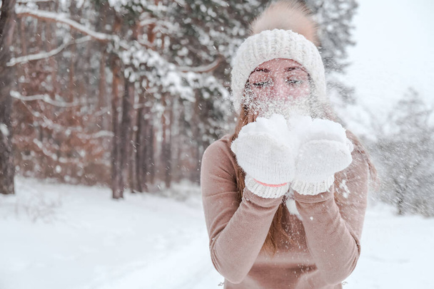 Noel, tatil ve sezon konsepti. Genç mutlu kadın kış ormanlarında kar üflüyor. Sıcak giysi, eldiven ve şapka.. - Fotoğraf, Görsel