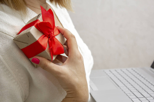A woman's hand holds a gift box with a red bow and a laptop in her hands. The concept of online shopping for holiday gifts. Give gifts online via the Internet - 写真・画像