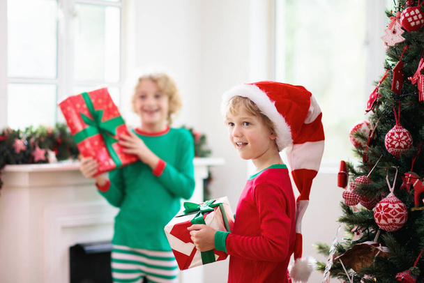 Παιδιά στο χριστουγεννιάτικο δέντρο και τζάκι την παραμονή των Χριστουγέννων. Οικογένεια με παιδιά που γιορτάζουν τα Χριστούγεννα στο σπίτι. Αγόρι και κορίτσι με ασορτί πιτζάμες στολίζουν χριστουγεννιάτικο δέντρο και ανοίγουν δώρα. Χριστουγεννιάτικα δώρα για το παιδί. - Φωτογραφία, εικόνα