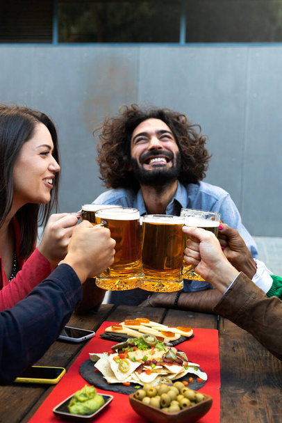 Šťastný a usměvavý běloch, který si připíjí s přáteli s pivem v baru venku. Slavnostní přípitek. Svislý obrázek. - Fotografie, Obrázek