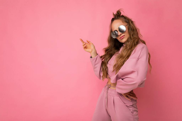 Photo shot of young beautiful attractive cute cool nice curly blonde woman wearing trendy and stylish pink sport suit and sunglasses isolated over pink background with copy space for text - Foto, Imagem
