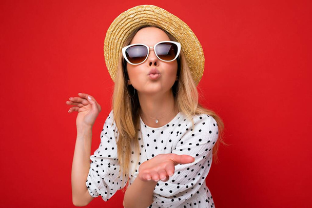 Foto-Foto von jungen schönen niedlichen glücklich blonde Frau trägt lässiges Kleid, Sonnenbrille und Strohhut isoliert über rotem Hintergrund mit Kopierraum und geben Kuss - Foto, Bild