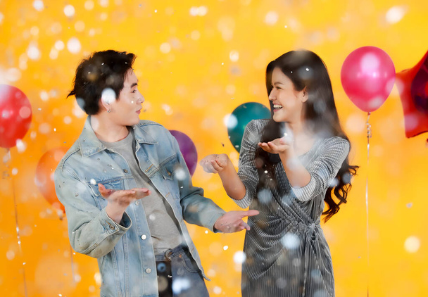 Studio shot of Asian happy young male and female lover couple model in fashionable wears play andi fun withpaper confetti in celebration party with balloons on yellow background. - Photo, Image