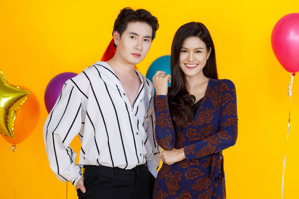 Portrait studio shot of Asian male and female teenager lover couple model in fashionable wears standing posing smiling look at camera in front colorful round and star balloons on yellow background. - Photo, Image