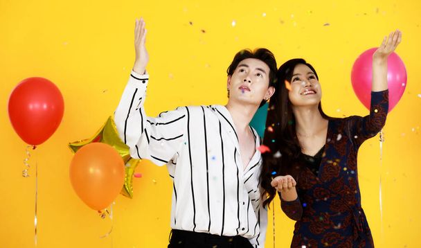 Studio shot of Asian happy young male and female lover couple model in fashionable wears playing fun with shiny paper confetti in celebration party with balloons on yellow background. - Photo, Image